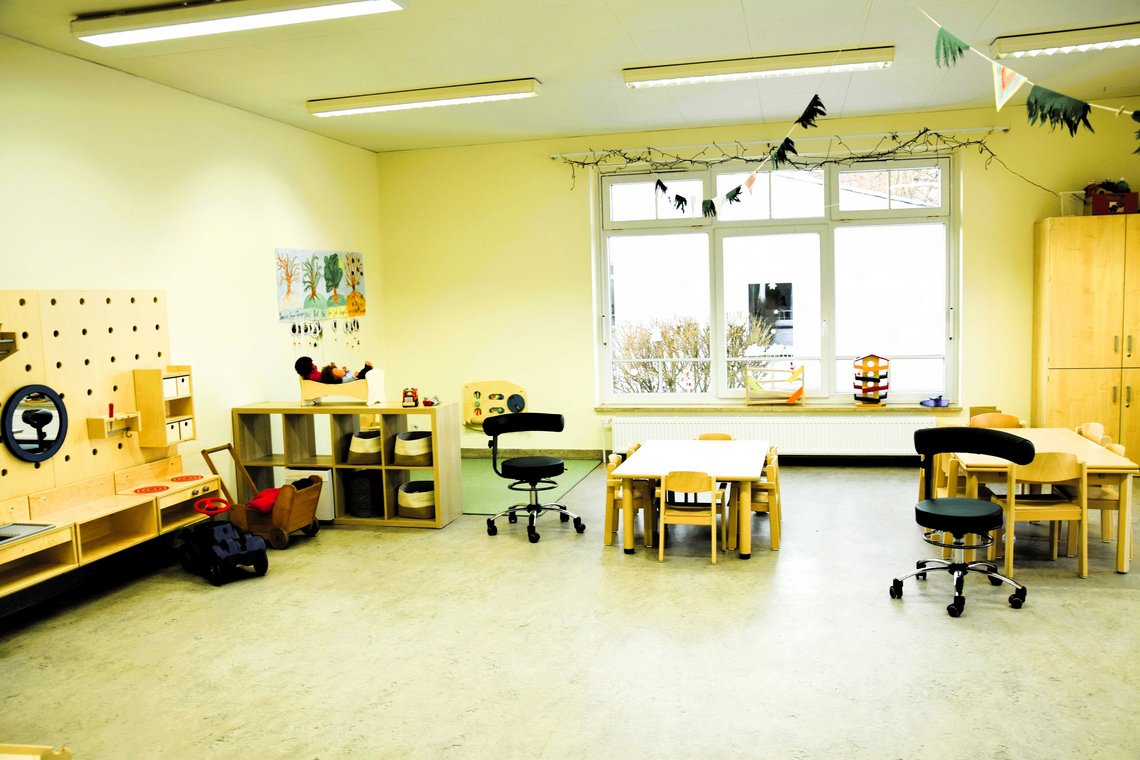 Blick in den Gruppenraum mit Tischen und Stühlen sowie einer Spielküche links an der Wand